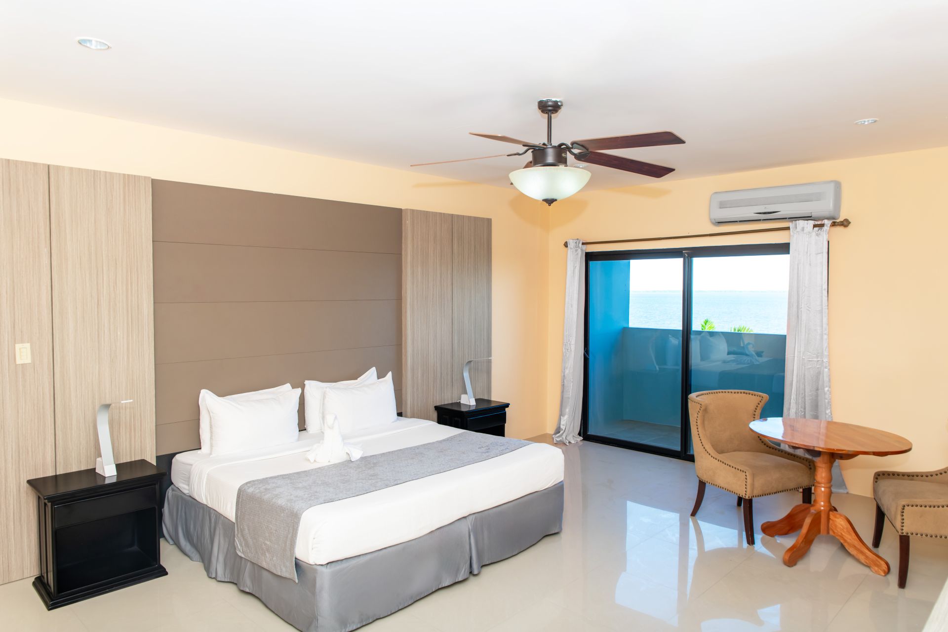 A hotel room featuring a king bed with white linens and a grey blanket, dark wooden nightstands with lamps, ceiling fan, small round table with two chairs, and a balcony with sliding glass doors offering a view of the outdoors.