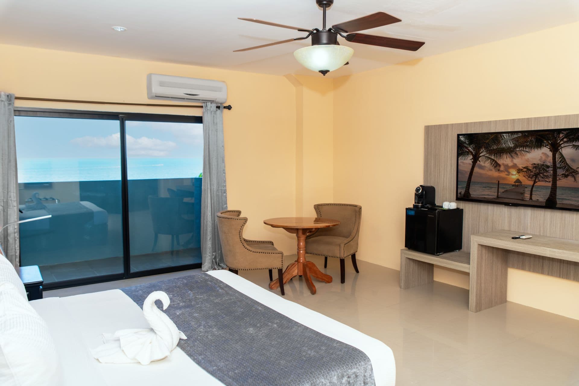 A well-lit hotel room with a balcony providing natural light. The room includes a king bed, a small round table with two chairs, ceiling fan, and flat-screen television.