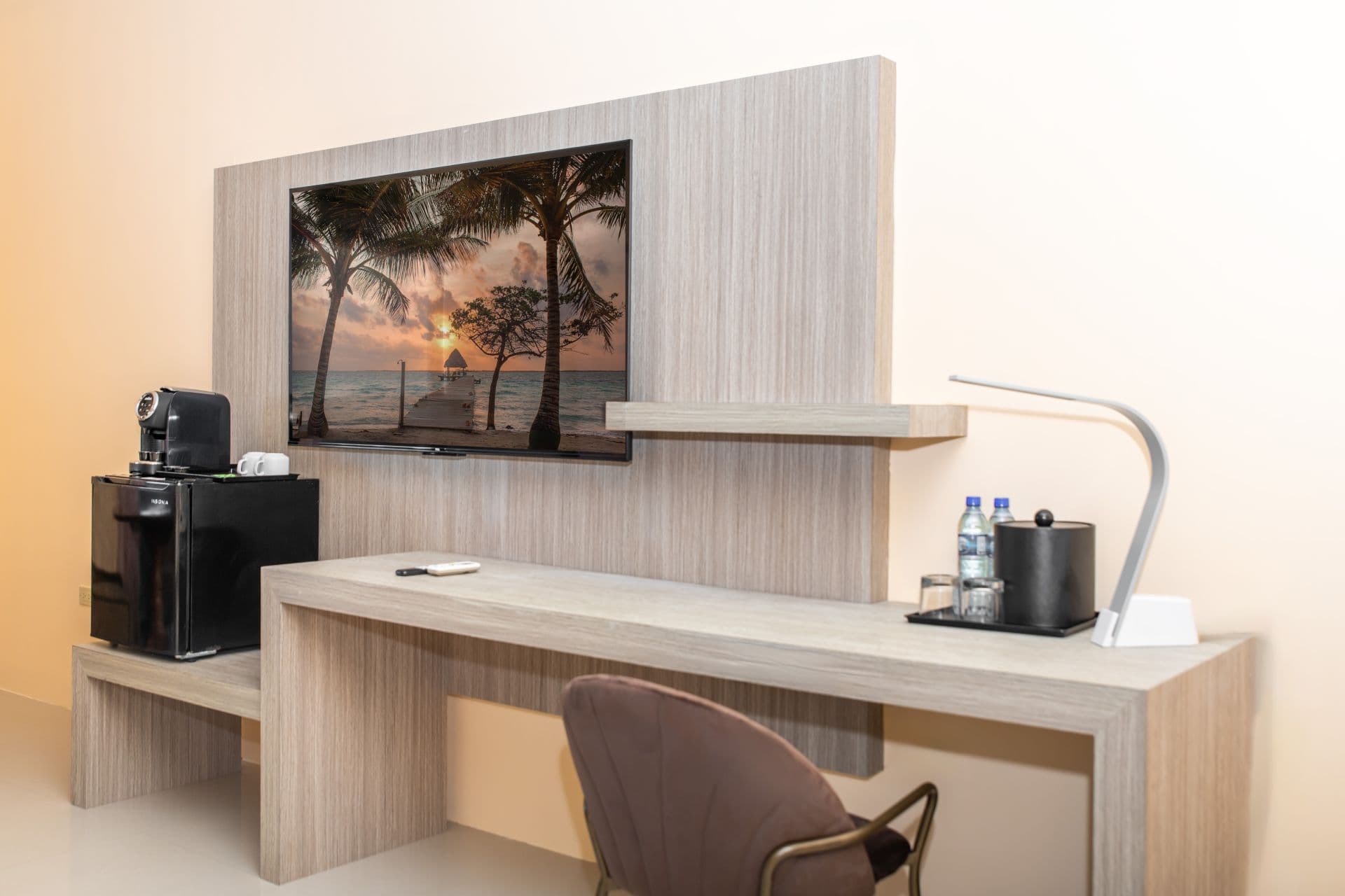 Desk area featuring a wall-mounted flat-screen television displaying a tropical scene, a black coffee maker with a water dispenser, a modern desk lamp, and a chair partially visible underneath the light-colored desk.