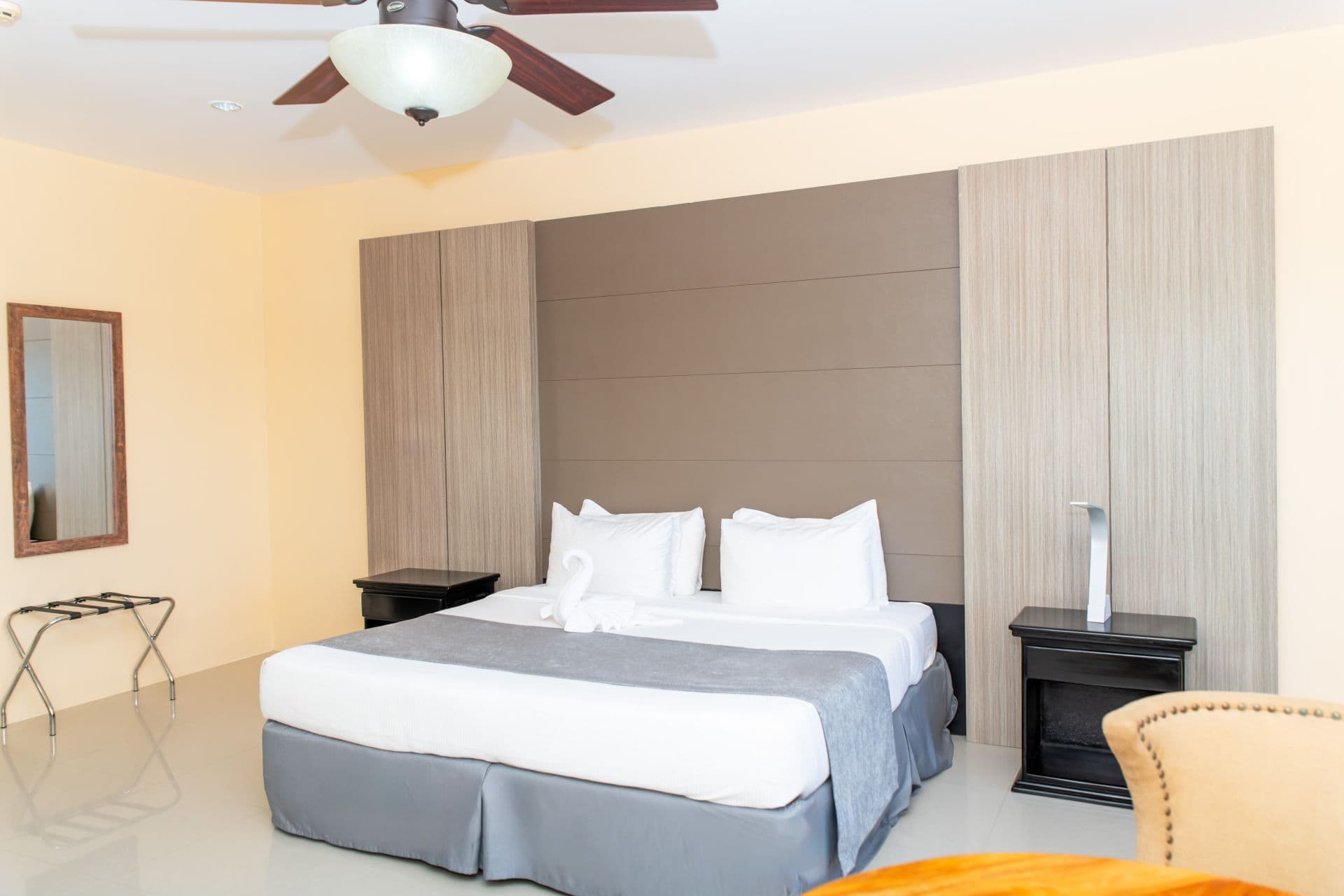 Hotel room featuring a king bed with white linens and a grey blanket, flanked by dark wooden nightstands with lamps. A ceiling fan with a light fixture is visible on the ceiling. A mirror and luggage rack are in the background.