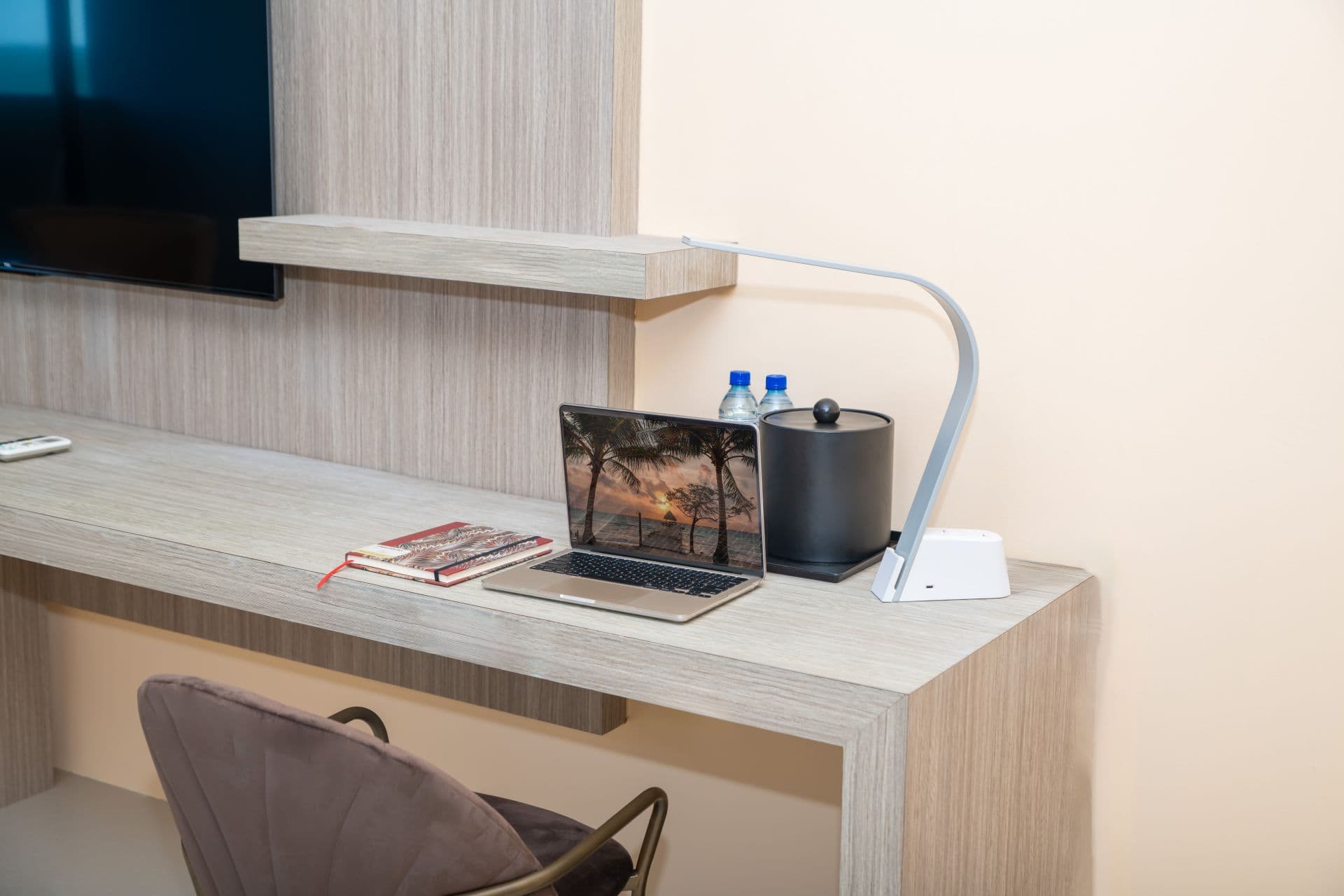 A close-up of a light-colored desk with a laptop computer displaying an image, a flexible desk lamp illuminating the workspace, two water bottles in a black container, and a notebook. A chair is partially visible underneath the desk. A portion of a wall-mounted television is visible in the upper left.