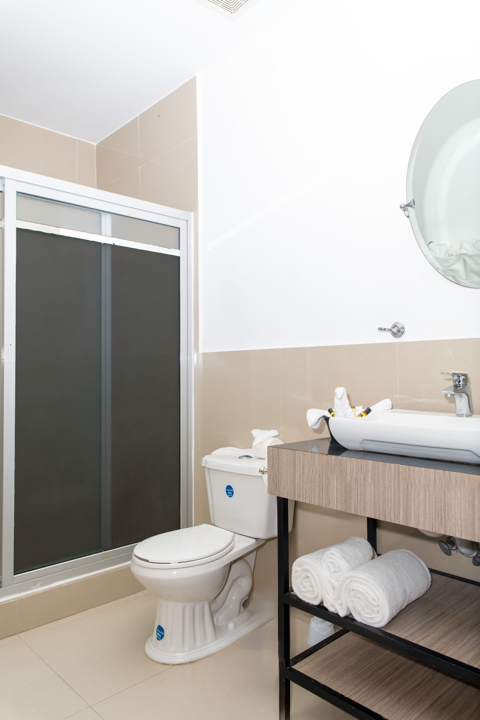 Interior view of a bathroom featuring a white toilet, a modern sink with a wooden vanity and a shelf holding white towels, and a shower with sliding glass doors. A round mirror is visible above the sink. The walls are painted in neutral tones.