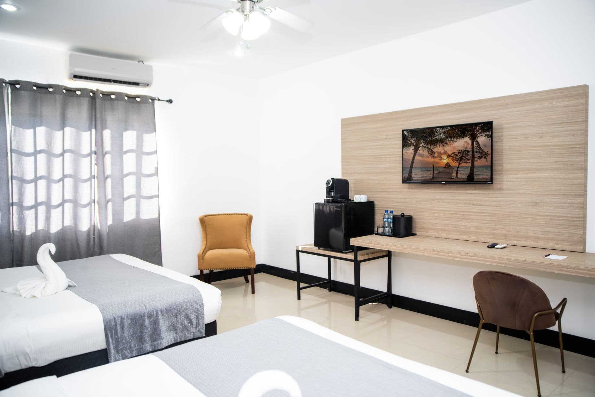 A hotel room designed with two beds, a dedicated workspace with a desk and chair, and an entertainment area featuring a television. A window with curtains is on the left side of the room.