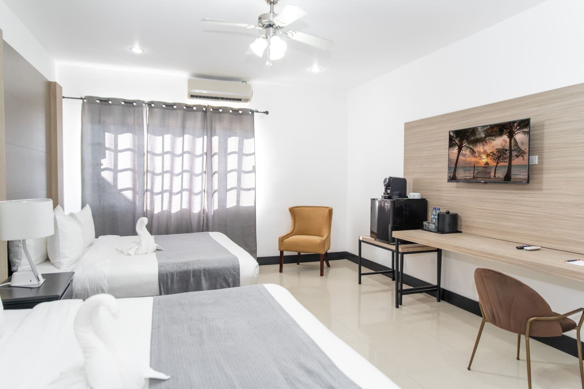 A well-appointed hotel room offering two beds, a dedicated workspace with a desk and chair, and an entertainment setup with a television. The room is lit by natural light from a window with curtains and has a ceiling fan for comfort.