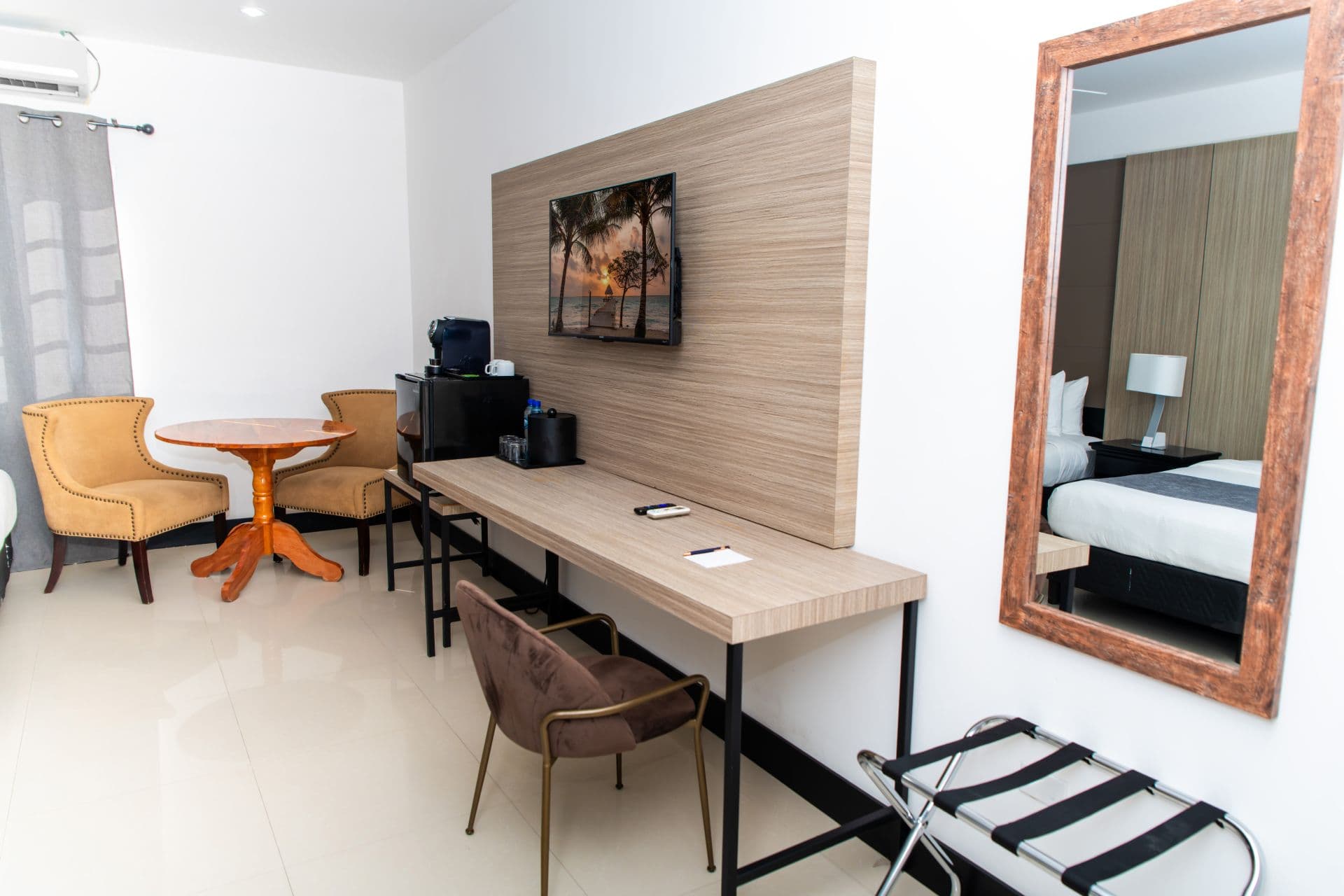 A hotel room with a desk and seating area. A prominent wooden-framed mirror reflects a view into another part of the room, showing a bed with white and grey linens.