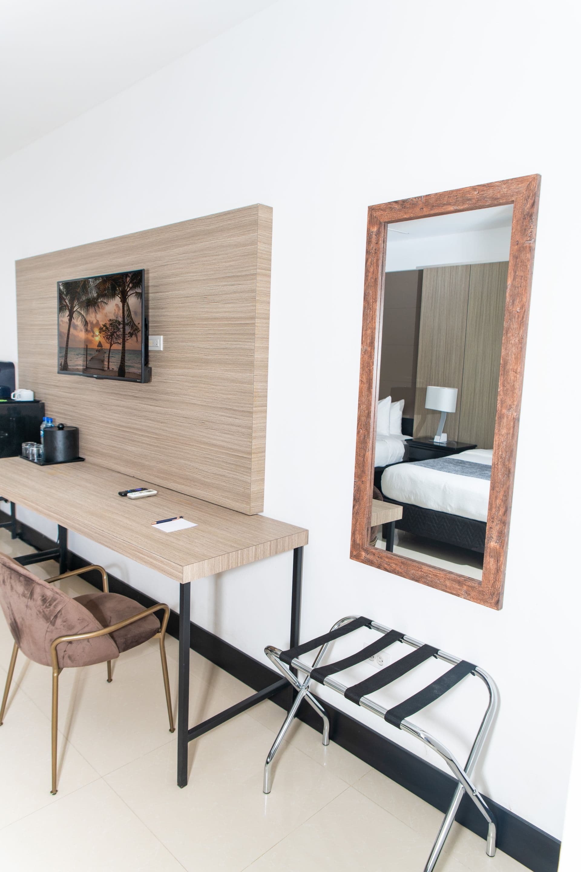 A functional workspace in a hotel room with a desk and chairs, providing an area for work or study. A television offers entertainment, and a mirror reflects the sleeping area with two beds. A luggage rack is available for convenience.