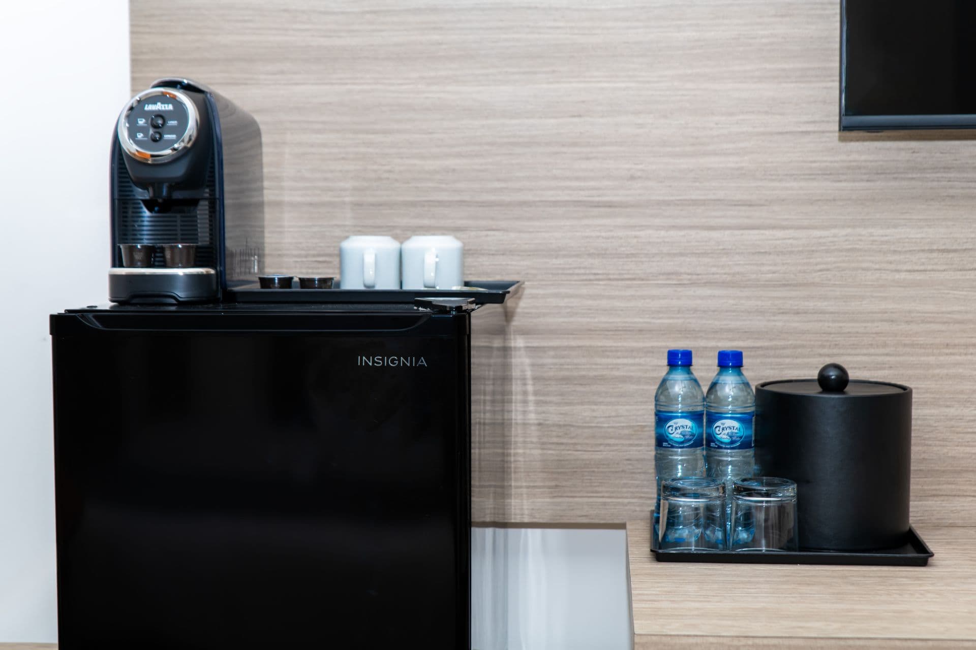 Hotel room amenities including a black mini fridge, a coffee maker for single-serve espresso, bottled water, drinking glasses, and an ice bucket, arranged on a countertop.