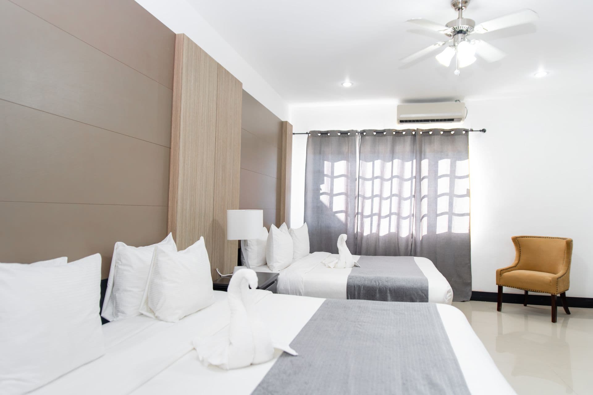 A bright and airy bedroom with two beds, upholstered chair, sunny window, and ceiling fan.