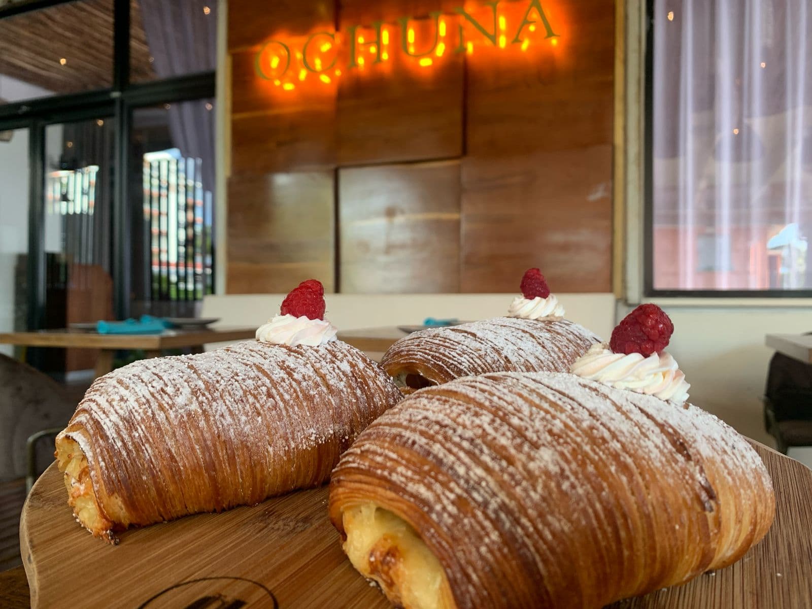 Three pastries topped with whipped cream and raspberries sit on a wooden platter in a cafe.