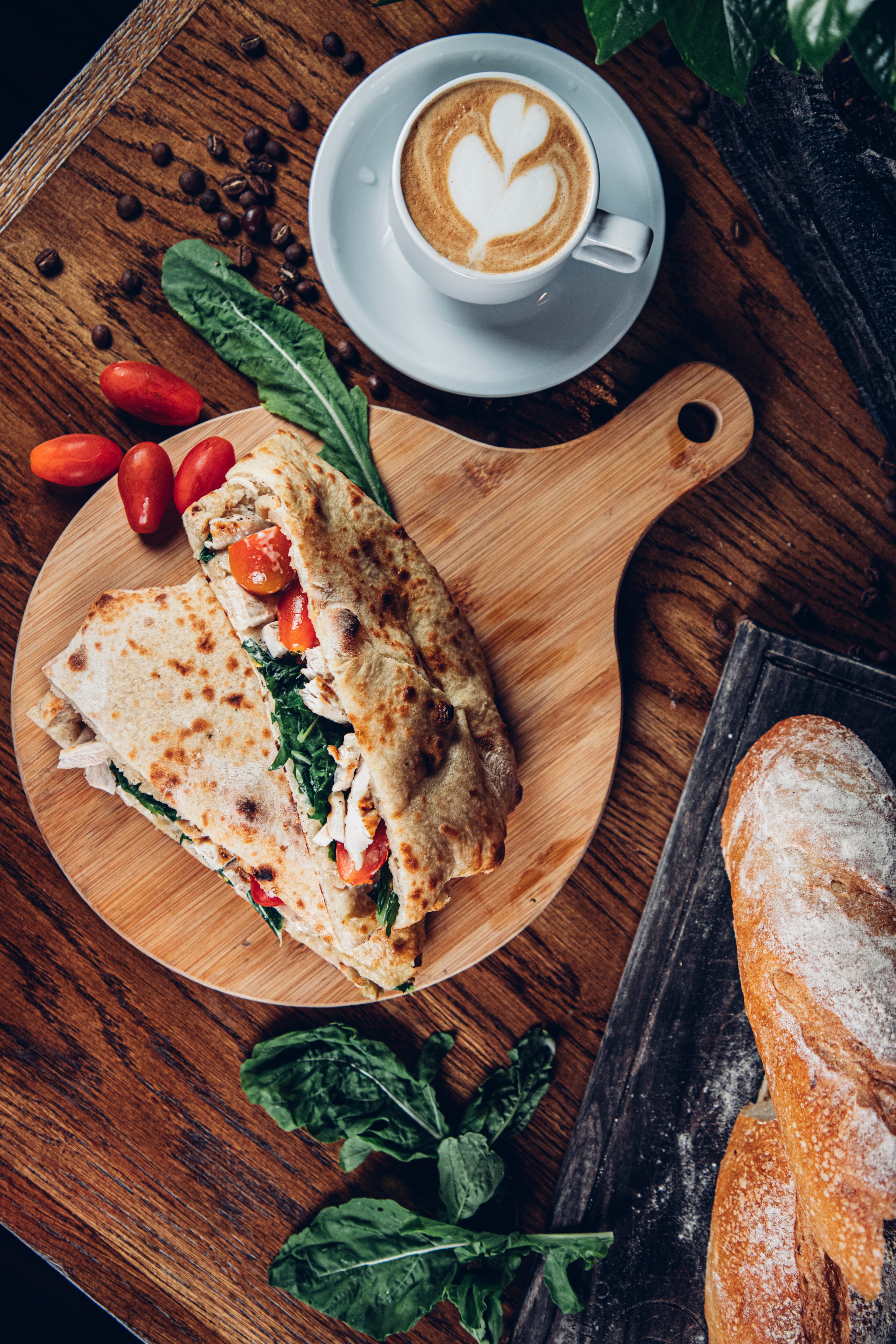 A wooden board displays a folded sandwich with chicken, tomatoes, and spinach alongside a cup of latte art and scattered coffee beans.