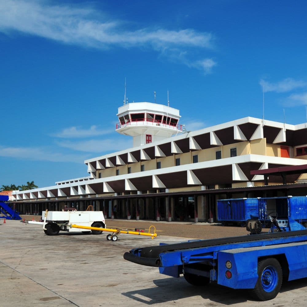 A bright blue sky above an airport terminal featuring a control tower and service vehicles on the ramp.
