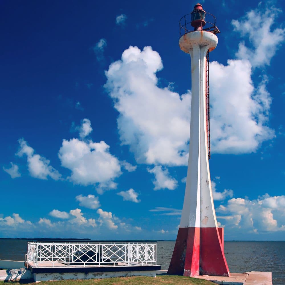A tall, white and red lighthouse stands next to a wooden dock under a blue sky with fluffy clouds.