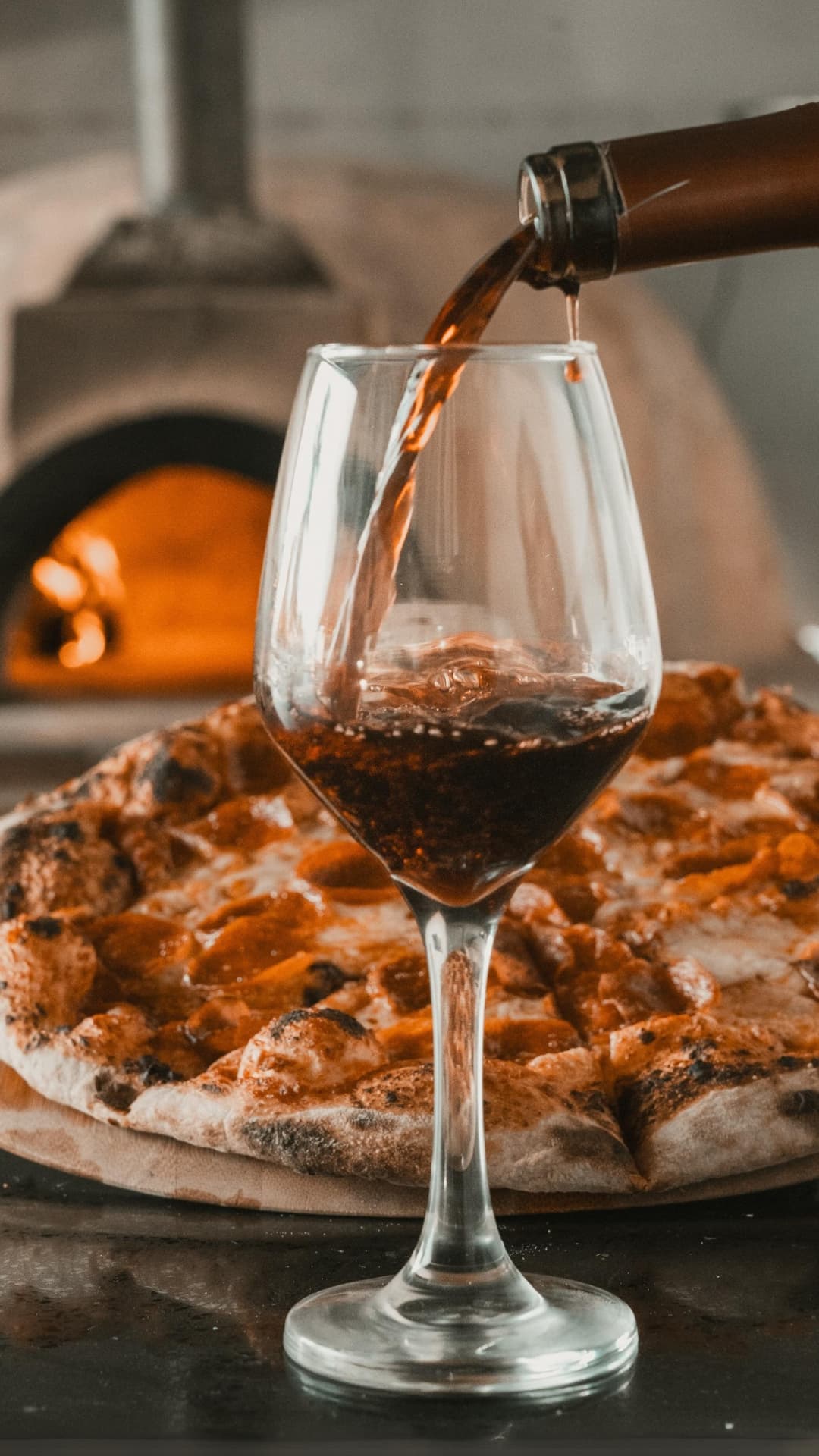 A glass is being filled with dark beverage in front of a freshly baked pizza.