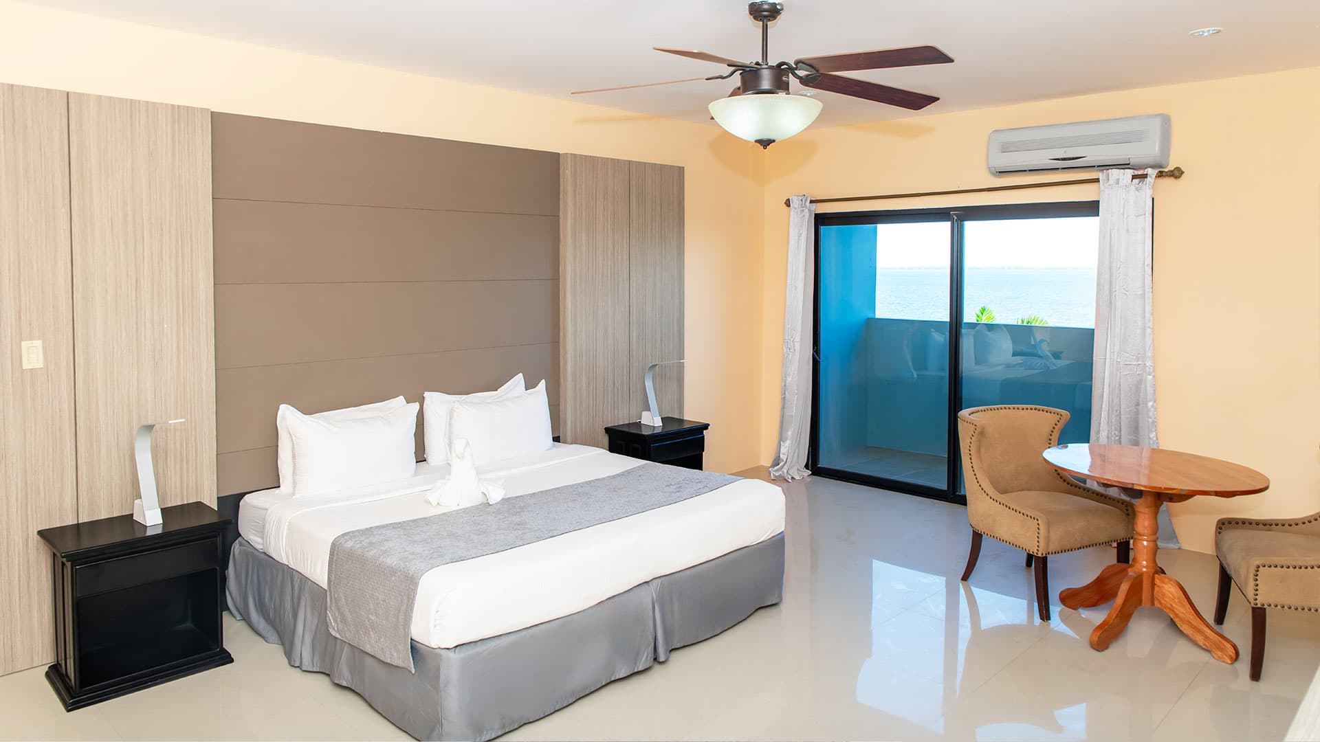 A modern hotel room featuring a bed, side tables, a small round table, and large windows with a sea view.