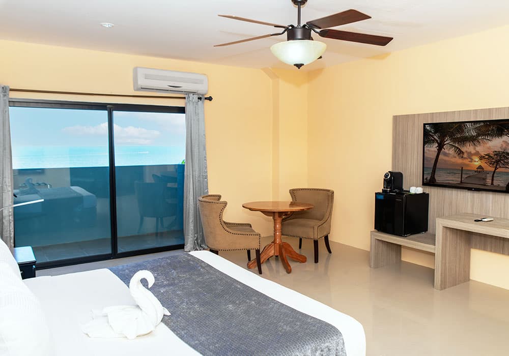 A bright hotel room featuring a bed with a towel swan, a dining table, a TV, and a scenic ocean view through large glass doors.