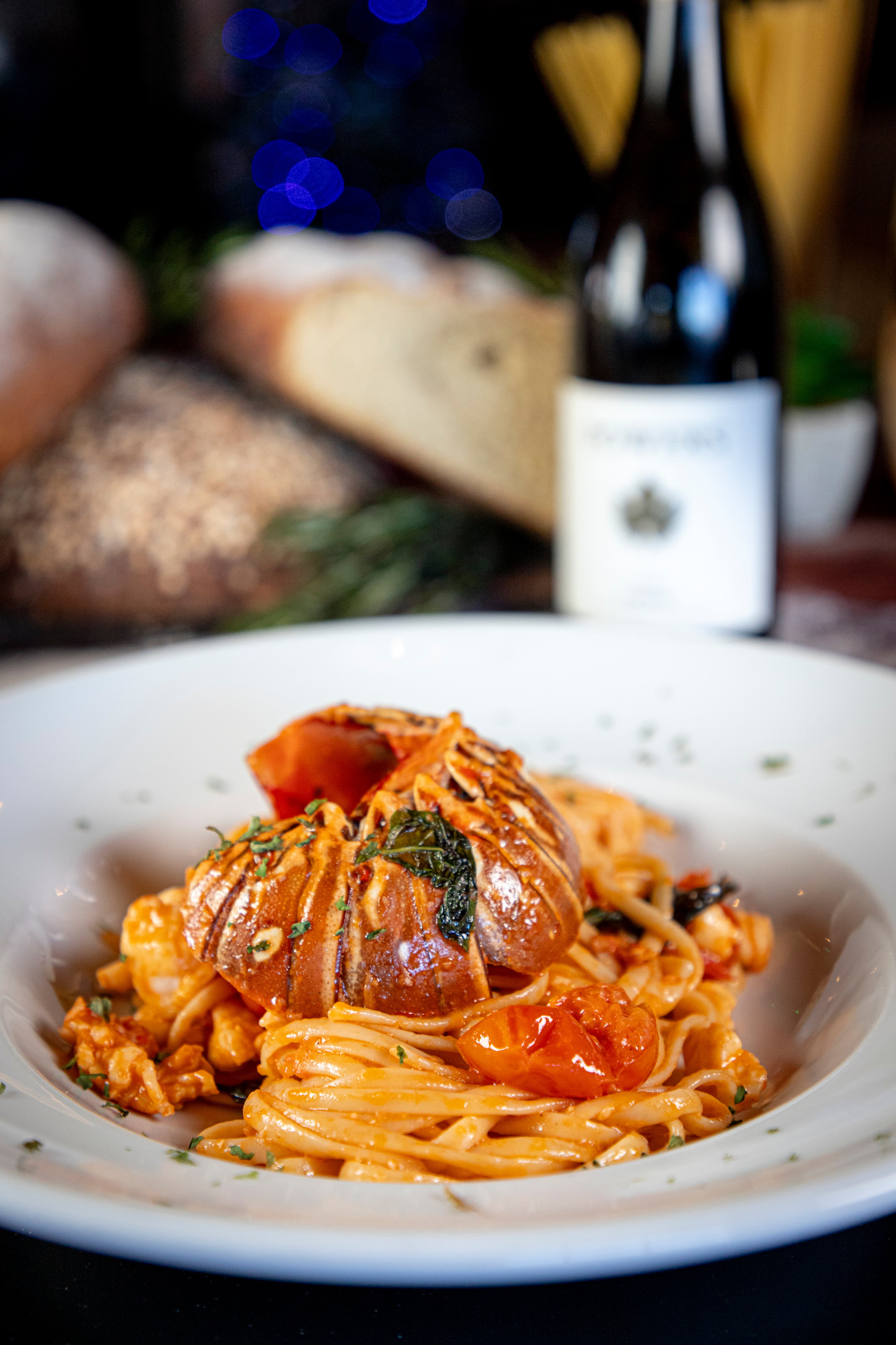A plate of spaghetti topped with lobster, cherry tomatoes, and herbs, with bread and a bottle of wine in the background.