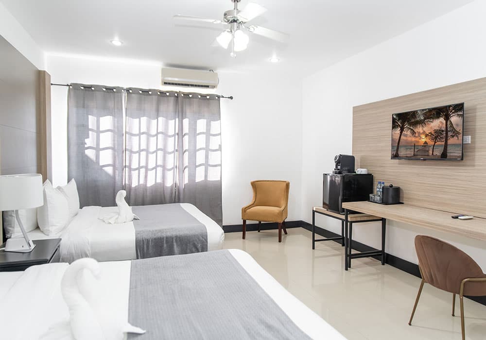 A modern hotel room featuring two beds, a work desk, and a TV, with light curtains allowing natural light.