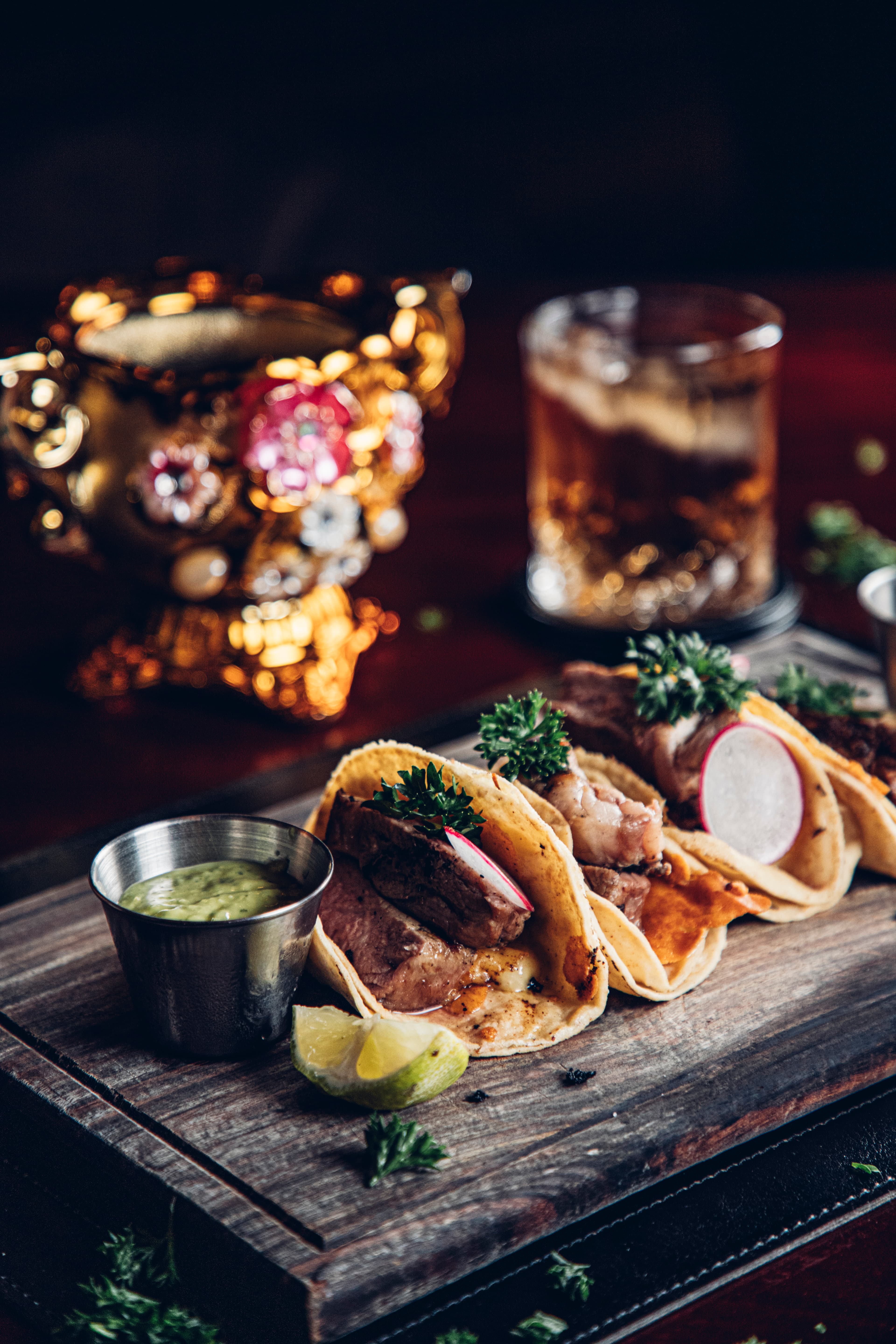 Three tacos filled with meat and garnished with parsley and radish, served with lime and green sauce.