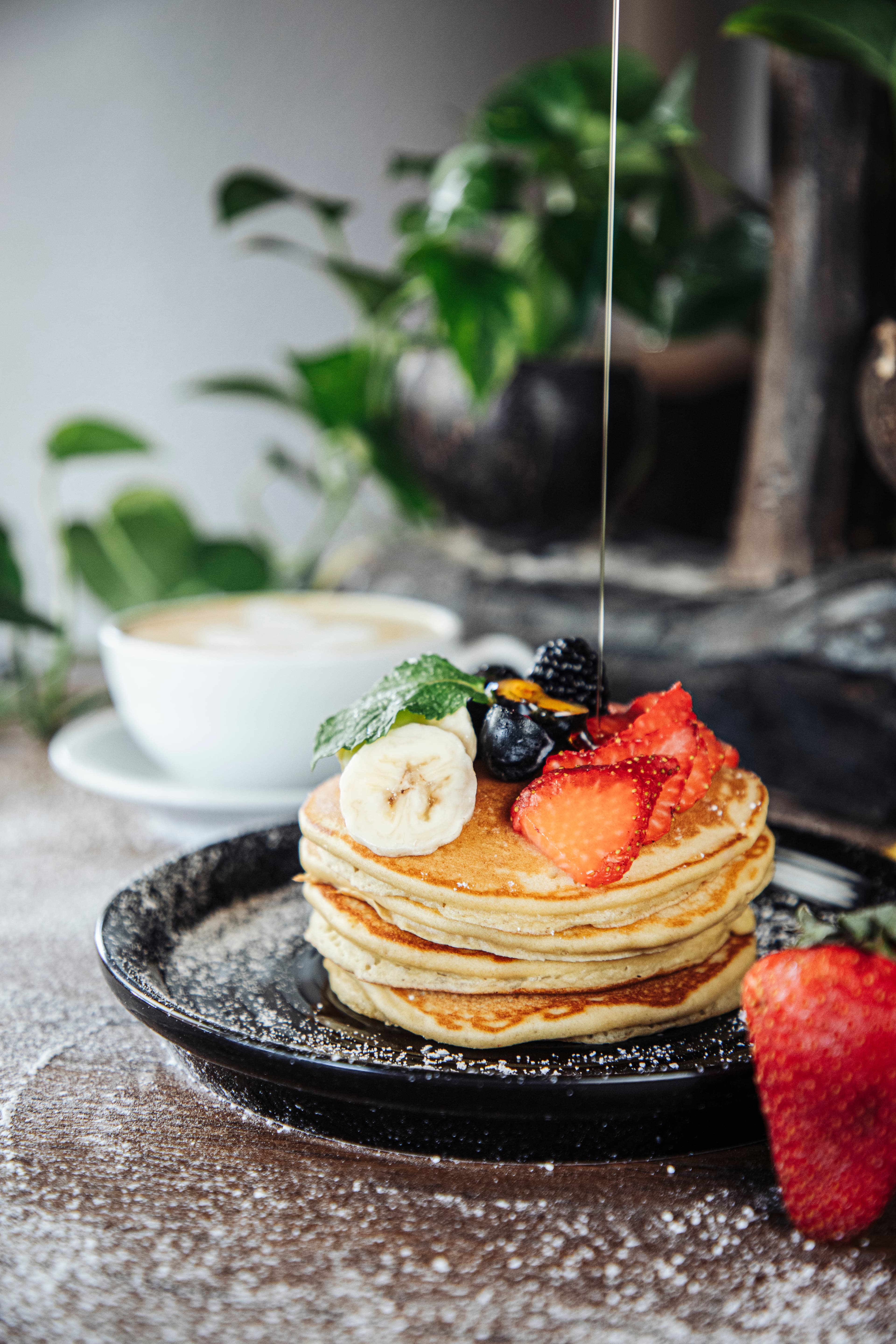 A stack of pancakes topped with sliced bananas, strawberries, blueberries, and mint, with syrup drizzling down, alongside a cup of coffee.